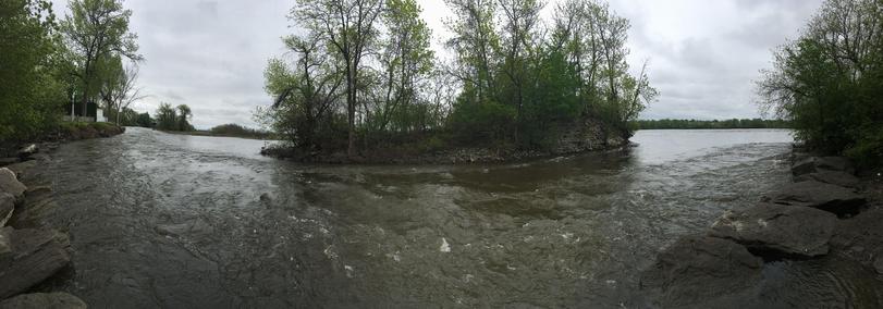 Panoramic photo of the Prairies River. On the left and right, there are green trees. In the middle, the river flows around a small island. That island has green trees as well. It is cloudy. 
- - -
Photo panoramique de la rivière des Prairies. À gauche et à droite, il y a des arbres verts. Au milieu, la rivière coule autour d'une petite île. Cette île a aussi des arbres verts. C'est nuageux.