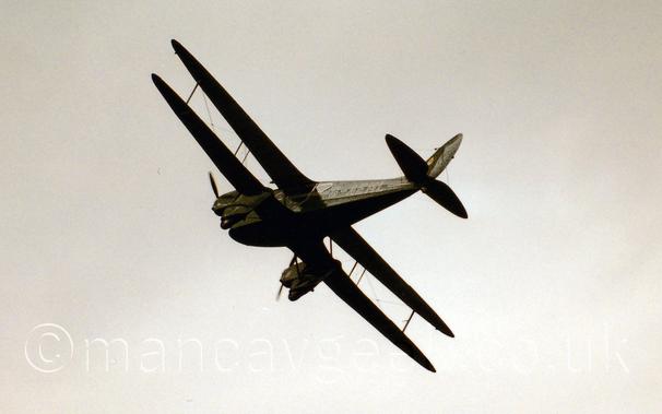 Low side view of a green, twin engined bi-plane airliner flying from right to left at a low altitude, banking to the right and flying away from the camera.
The registration "G-AEML" is on the side of the rear fuselage, outlined in white.
Flat white sky fills the rest of the frame.