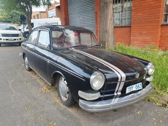 A black VW Type 3 fastback with silver racing stripe, sitting at the side of the road in an industrial estate. The tyres are soft, the interior dirty.  It looks as though it has not moved for months