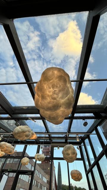 Foto aus einem Restaurant, das rundum durch Fenster den Blick nach außen ermöglicht. Zu sehen sind Wolken, die von der Decke hängen, dahinter echte Wolken am blauen Himmel. Im Hintergrund ist das geschichtsträchtigen Gelände der ehemaligen Zeche Nordstern in Gelsenkirchen-Horst zu erkennen.

Photo of a restaurant that allows the view to the outside all around through the window. You can see clouds hanging from the ceiling, behind them real clouds in the blue sky. In the background, the historic site of the former Nordstern mine in Gelsenkirchen-Horst can be seen.