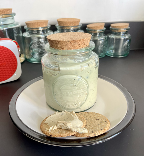 photo of two oatcakes (one of which is covered with a creamy cashew cheese) on a cream-coloured plate that has a black rim. behind the oatcakes is a heavy glass jar filled with cashew cheese & sealed with a cork lid. in the background is a portable speaker & a row of glass jars.