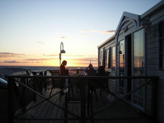 Rechts ein Mobilhome mit Fensterfront, der Blick geht über die Terrasse davor aufs offene Meer. Die Sonne erleuchtet den Horizont, die Farben verschwimmen in grau, blau und orange. Die Silhouette von Kindern im Bild.
