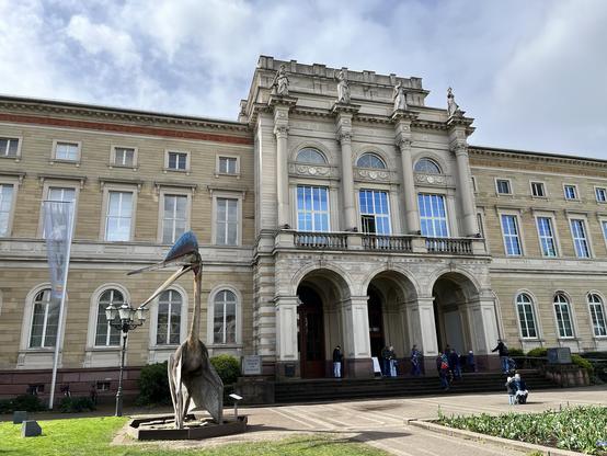 Ein imposantes Gebäude mit großen Bögen und Säulen ist zu sehen. Im Vordergrund ist die Skulptur eines Flugsauriers zu sehen. Links ist ein Banner zu sehen. Man sieht Menschen vorm Gebäude und viel Grün und Blumenbeete. Der Himmel ist mit Wolken durchzogen.