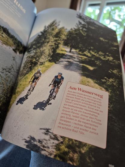Two cyclists ride along a gravel path surrounded by lush greenery. They wear helmets and cycling gear, enjoying a bright and sunny day. A German-language information board titled "Am Wasserweg" details the cycling route, mentioning landmarks like the Isar River and Bad Tölz. Trees frame the background, with glimpses of the river enhancing the scenic setting.