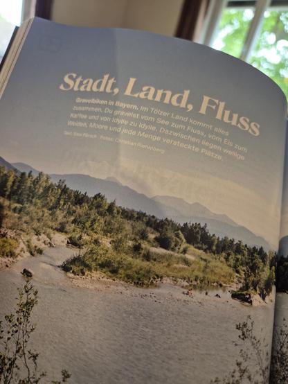 A  book lies open, showing a page with a full-page color photograph of a misty mountain range with dense green forests. The opposite page features German text, formatted in a clean and elegant font, introducing a nature-themed topic. The lighting in the image is soft and natural, emphasizing the textures of the book and the wooden background.