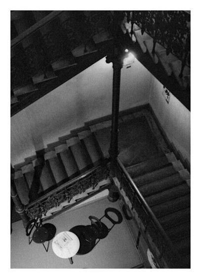 View down a staircase, it's an old wide staircase with lots of art details. Down at the ground floor is a little round white bistro table with a marble top, two black chairs around it throwing deep black shadows.