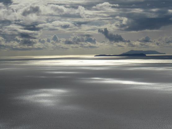 View from a vantage point west into the sun over the Ionian Sea. Mottled cloud sends reflections over the flat sea.  A couple of islands lie on the horizon.