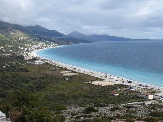 View from a hillside down onto the yellow arc of a long beach beside turqoise water. Small resorts are scattered amongst olive groves.