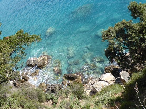 View down from above about 10 metres down a steep cliff to a small bit of rocky coastline. The water is a vivid turqoise colour.