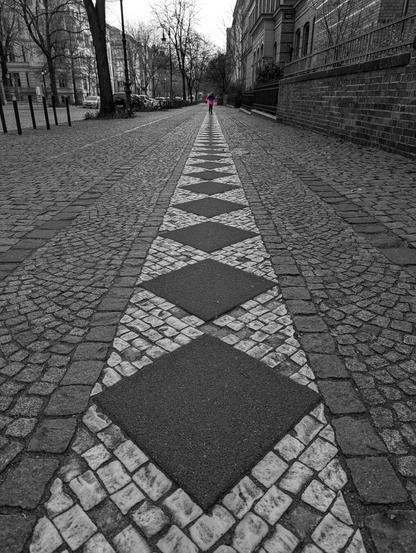 The image is a black and white photograph of a cobblestone pavement next to a road (visible on the left). The pavement stretches into the distance, creating a strong sense of perspective. A child wearing a bright pink winter jacket is running towards the viewer. The cobblestones on the pavement are arranged in a pattern, with a central strip of diamond shapes running along its centre. Buildings line the right side of the pavement, and bare trees are visible on the left, next to the road.