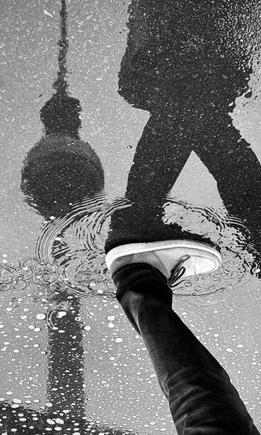 A black and white image of a person's foot stepping into a puddle, with the reflection of a tall structure, likely a tower, visible in the water. Raindrops create ripples around the foot.