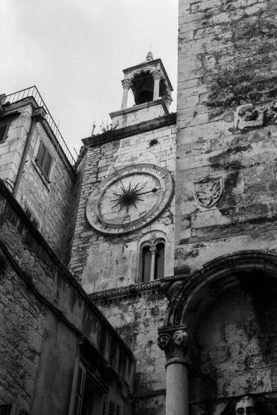 The image captures a couple standing in front of the historic Clock Tower in Split, Croatia. In the foreground, an archway and a column add depth to the composition. The grand stone buildings serve as a backdrop, emphasizing the location's historical significance.