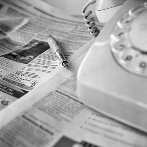 A high-angle, black and white close-up shot shows an old-fashioned rotary phone and a pen resting on an open newspaper. The telephone, positioned on the right side of the frame and slightly out of focus, has a coiled cord connecting its receiver to the base. The rotary dial, with numbers and letters, is partially visible. To the left, an open newspaper is spread out, its headlines and text legible in parts. A metallic pen with a cap rests on the newspaper, pointing towards the top left. The focus is sharpest on the pen and the newspaper in the foreground, with the background slightly blurred.