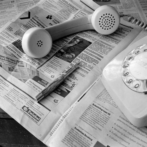 A high-angle, black and white close-up shot captures an old-fashioned rotary phone and a pen resting on an open newspaper. The telephone's detached receiver lies horizontally across the upper part of the newspaper, with the earpiece and mouthpiece facing slightly upwards. The rotary phone base is visible on the right side of the frame, its dial with numbers and letters partially in view. A pen, possibly with a wooden or textured grip, rests on the newspaper below the receiver, pointing towards the bottom left. The newspaper is spread open, displaying various German headlines, advertisements, and text, with some legible phrases and a phone number. The scene suggests a moment of contemplation or a break from reading and communication.
