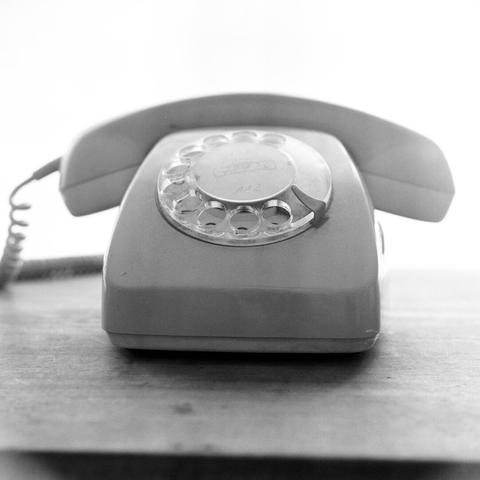 A black and white, eye-level shot captures an old-fashioned rotary telephone resting on a wooden surface, set against a bright, overexposed background. The telephone, a classic model from possibly the 1960s, is positioned centrally in the frame. Its receiver rests on top of the base, and a coiled cord extends from the left side. The clear plastic rotary dial with finger holes and numbers is clearly visible on the front of the phone. The wooden surface below the phone shows some faint grain or texture. The bright background creates a high-key effect, silhouetting the phone and emphasizing its form.