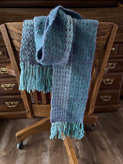 A wooden desk and chair provide backdrop for a scarf draped over the chair. It is two tones of turquoise along the length and woven throughout is purple making a sorta floral like pattern. The backside it looks different, more diamond shaped with dots. While the fringe appears different lengths that is just perspective, sorry! Haha.