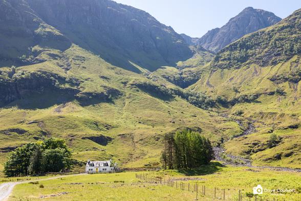 A photograph of a white cottage at the foot of some stunning hills in the valley of Glencoe. Shot early afternoon on a bright sunny day.