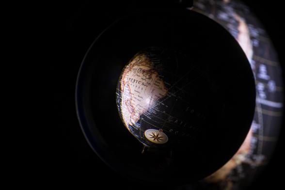 Das Bild zeigt einen beleuchteten Globus durch den Blick einer Glaskugel im Dunkeln, der Südamerika hervorhebt.

Das Bild ist stark von Dunkelheit dominiert, mit einem einzigen Lichtpunkt, der auf einen kleinen, antiken Globus fällt. Der Globus ist dunkel gefärbt, mit antiquierter Kartografie, die Kontinente und Ozeane in verwaschenen, erdigen Tönen zeigt. Südamerika ist deutlich beleuchtet, während der Rest der Welt in Schatten liegt. Der Globus selbst scheint auf einem Standfuß zu stehen, der jedoch im Dunkeln kaum zu erkennen ist. Im Hintergrund ist ein verschwommener, ebenfalls dunkler Globusausschnitt zu sehen, wodurch ein räumlicher Tiefeeffekt entsteht. Die Gesamtatmosphäre ist geheimnisvoll und nachdenklich, fast meditativ. Die Beleuchtung des Globus schafft einen Fokus auf Südamerika, der eine symbolische Bedeutung haben könnte (z.B. Reise, Exploration, Entdeckung eines bestimmten Ortes). Der Kontrast zwischen Licht und Dunkelheit betont die zentrale Rolle des Globus im Bild und erzeugt ein Gefühl von Konzentration und Introspektion.