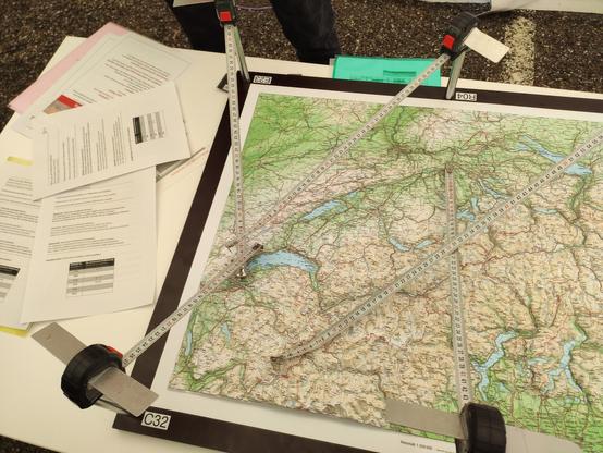 Ein Modell, welches die Funktionsweise von GNSS demonstriert: Um eine Reliefkarte der Schweiz sind auf ca 20cm hohen Stäben ausrollbare Massbänder drehbar montiert. Diese Massbänder symbolisieren die Satelliten. Nun ist die Aufgabe, durch Abrollen der Massbänder auf bestimmte vorgegebene Abstände (welche die Signallaufzeiten darstellen) den Punkt auf der Karte zu finden, zu dem alle Abstände passen.