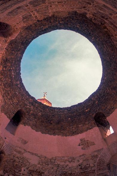 This is a view from inside a circular stone ruin. Looking up through an oculus at the blue sky, a church cross is visible above the wall.
