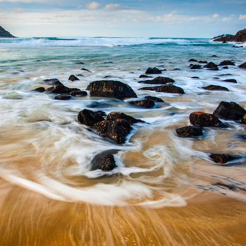 Rocks on a golden sand beach surrounded by swirly waves caused by using a slow shutter speed on the camera. The open sea and waves are in the distance.