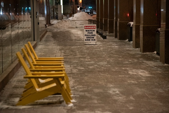 Another, wider and more modern colonnade, this one with a line of four yellow muskoka chairs and salt stains on the paving bricks. A sandwich sign reads “DANGER: watch for falling snow and ice”.