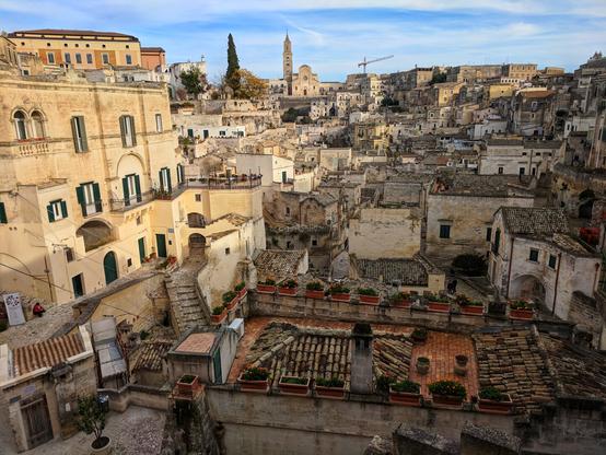 The image captures a panoramic view of Sassi di Matera from Belvedere Luigi Guerricchio in Matera, Italy. The scene is dominated by the ancient cave dwellings and buildings, constructed from pale, honey-coloured stone, densely packed together across the hillside. Many of the buildings feature dark green shutters. Red flower boxes line the rooftops. In the background, a church with a tall spire rises above the town. The sky is a mix of blue and wispy white clouds.