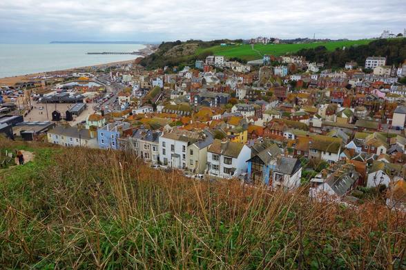 The image shows a panoramic view of Hastings, a town on the south coast of England. The view is taken from a slightly elevated position, with grassy slopes in the foreground. The town stretches along the coastline, with a mix of residential and commercial buildings. The buildings are mostly low-rise, with a variety of architectural styles and colours. The rooftops are a mix of red tile and slate. A long beach runs along the edge of the town, with a pier extending into the sea. The sea is a deep blue, and the sky is overcast with grey clouds. The image captures the essence of a traditional British seaside town.