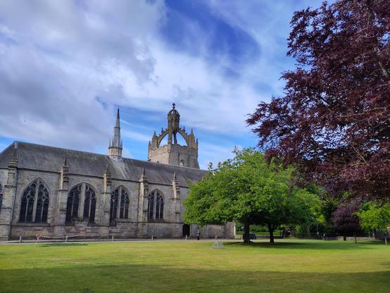 A sunny view of King’s College at Aberdeen University, seen from just outside Elphinstone Hall. Some trees (one quite purple) are visible on the right. It's a little cloudy in the background despite the sun.