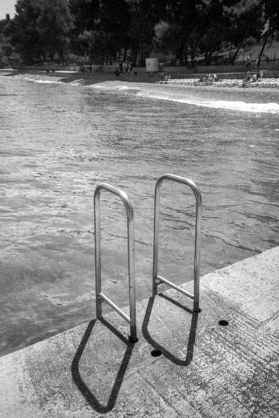 The scene unfolds at a beach in Croatia, where sunlight bathes the shore. In the background, adults and children relax by the water. Closer to the viewer, a metal ladder leads into the sea.