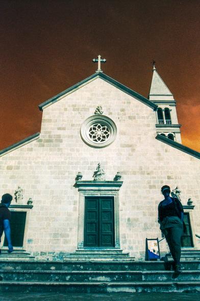 The image captures a scene at what appears to be the Church of Our Lady of Health in Supetar, Croatia. The edifice dominates the background, its weathered facade rising majestically against the sky. In the foreground, two figures are present: one on the left gazing towards the church and another on the right descending the steps in front of the church.