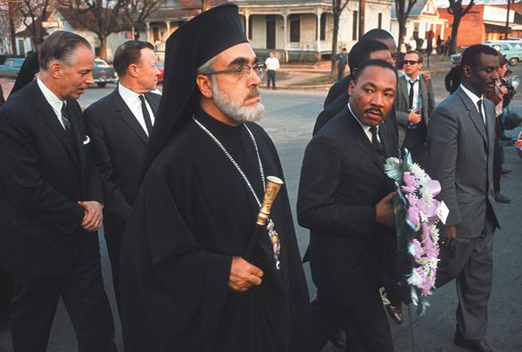 Dr. Martin Luther King Jr. walks alongside Archbishop Iakovos of the Greek Orthodox Church during the Selma civil rights march in 1965. Dr. King, dressed in a dark suit and holding a funeral wreath, gazes solemnly to his side amid a diverse group of clergy and supporters. Archbishop Iakovos, in his black clerical robes and kalimavkion, holds a staff and wears a serious, resolute expression.