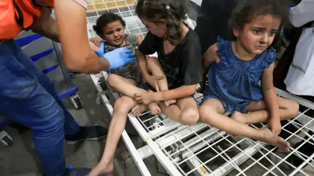 Wounded children receiving treatment in a hospital, three to a bare bed.