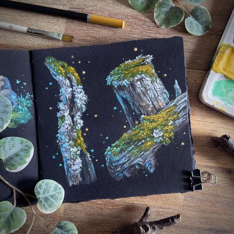 A photo of an open, square sketchbook with black paper, placed on a wooden table and surrounded by a palette, a paint brush, a coloured pencil, leaves, and branches for decoration. The sketchbook page features moss and lichen-covered branches and tree stumps with tiny glowing dust particles floating around them. The lichen is pale grey and the moss a warm green and orange colour.