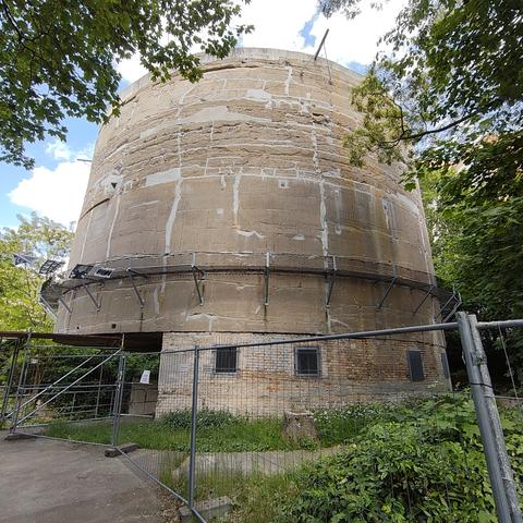 Umgeben von Bäumen und hinter einem Bauzaun steht der Schwerbelastumgskörper, ein mehrere Meter hoher Zylinder aus Beton.