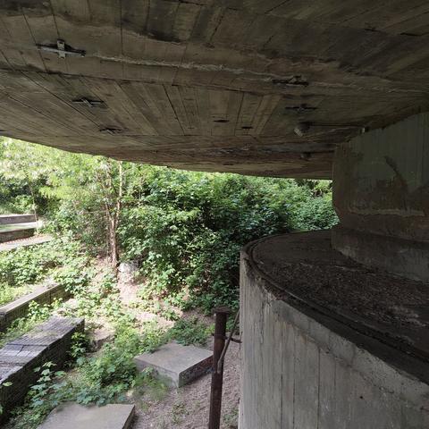 Blick auf die Unterseite des überstehenden oberen Teils des Schwerbelastungskörpers. Im Hintergrund grünes Gebüsch.