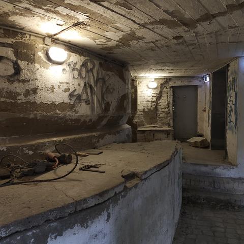 Zwei Lampen erhellen das Innere des Schwerbelastungskörpers. Von den Betonwänden und -decken Blätter weiße Farbe ab. Am Ende des Raumes ist eine graue Metalltür.