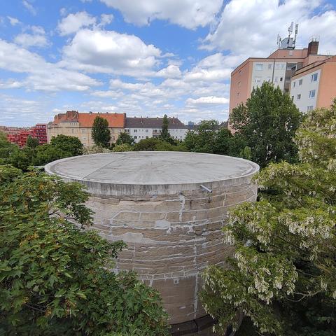 Blick von einer Aussichtsplattform auf die flache, runde Oberseite des Schwerbelastungskörpers, der von grünen Bäumen umgeben ist. Etwas weiter entfernt stehen einige mehrstöckige Wohnhäuser. Der Himmel ist heiter bis wolkig.