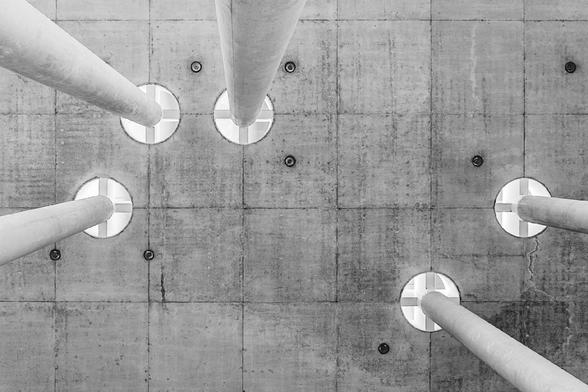 Black and white architectural shot taken from below, showing five cylindrical columns passing through round openings in a large concrete ceiling. The ceiling is made of square concrete tiles, and each circular opening is reinforced with a cross-brace structure.
