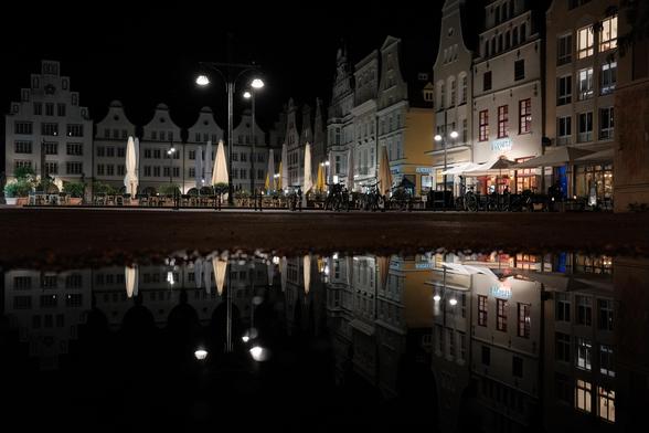 Das Bild zeigt die nächtliche Szenerie eines Platzes in einer Altstadt mit Restaurants, Terrassen, zusammengeklappten Sonnenschirmen und Fahrrädern vor einer großen Pfütze, in der sich alles spiegelt. Im Detail zeigt das Bild eine Reihe von Gebäuden im Stil einer europäischen Altstadt, die nachts beleuchtet sind. Die Gebäude sind mehrstöckig, hell und zeigen eine einheitliche Architektur mit Giebeln und vielen Fenstern. Einige der Fenster sind beleuchtet, was auf Aktivität in den Gebäuden hindeutet.  Auf dem Platz befinden sich die Außenterrassen mehrerer Restaurants mit Tischen, Stühlen und großen, hellen Sonnenschirmen, die zusammengeklappt sind. An den Terrassen stehen einige Fahrräder geparkt. Die Atmosphäre ist ruhig und friedlich, die Beleuchtung sorgt für eine warme und gemütliche Stimmung. Der Bildmittelpunkt wird durch eine Pfütze gebildet, die nahezu perfekt das Bild der Szenerie spiegelt.

The photo shows the night-time scene of a square in an old town with restaurants, terraces, folded up parasols and bicycles in front of a large puddle in which everything is reflected. In detail, the image shows a row of buildings in the style of a European old town, illuminated at night. The buildings are multi-storey, bright and have a uniform architecture with gables and many windows. Some of the windows are illuminated, indicating activity in the buildings.  On the square are the outdoor terraces of several restaurants with tables, chairs and large, bright parasols that are f