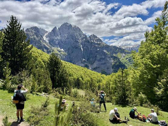 People sit in grass to rest or eat, with a mountain view framed by trees