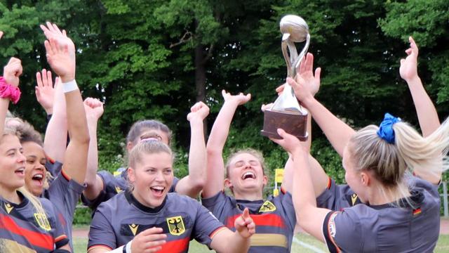 The German women's sevens rugby team celebrates with their first Bonn Sevens Cup at the Wasserland Bonn stadium. They raise the characteristic Bonn Sevens Cup. In the background are summery, green trees