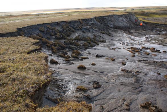 Photograph of a thawing permafrost area in Siberia. The landscape shows a steep erosion edge with slumped soil and peat blocks, as well as flowing mud patterns downhill. In the background, two people in bright clothing are examining the site. The surrounding area is flat and tundra-like, with sparse vegetation.