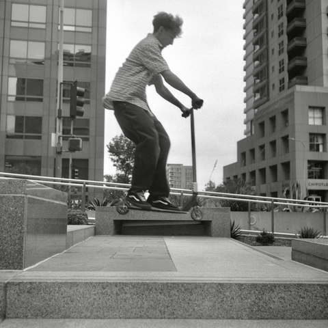 A black and white film photo. A shot of someone mid-flight as they jump steps with their scooter in downtown Los Angeles, California. Saturday, May 17, 2025.