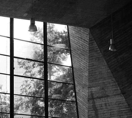 Black and white photograph showing a modernist interior with tall gridded windows revealing trees outside. Industrial pendant lights hang from the textured concrete ceiling with visible formwork patterns. Angular concrete walls create a dynamic geometric composition, highlighting the brutalist architectural aesthetic. ( made with help of Claude.ai)