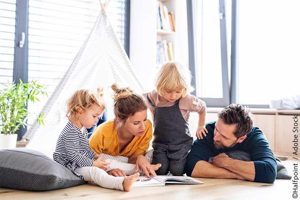 Familienzeit im Wohnzimmer, ein Kind zeigt auf ein Buch, während ein weiteres Kind, eine Frau und ein Mann aufmerksam daneben sitzen. Moderne Einrichtung mit hellem Tageslicht und pflanzlicher Dekoration.