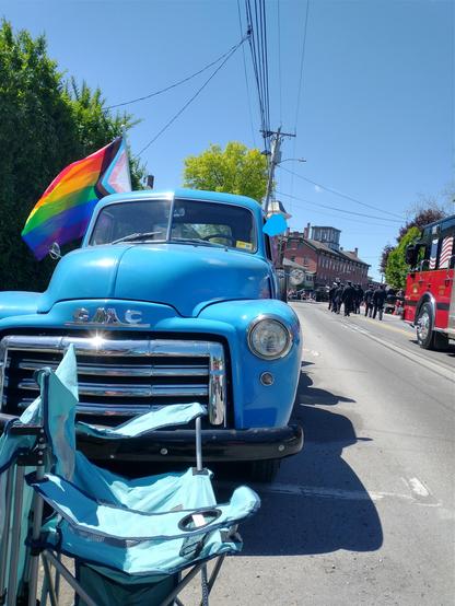 A 1950, powder-blue GMC truck, parked on the side of the street as a parade goes by. A large progress pride flag is flying from the truck.