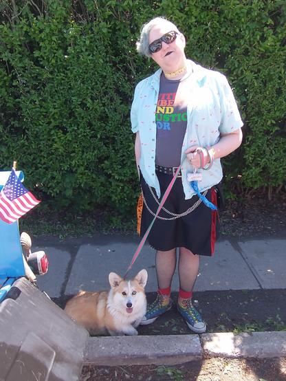 Me, standing on the sidewalk, next to the back bumper of the truck with my Corgi. I am wearing a pride kilt, rainbow pride Converse, a T-shirt that says "with liberty and justice for all" in rainbow colors, a camp shirt open over it that is light blue with pink flamingos, sunglasses, fishnets, a yellow flower choker and matching yellow flower bracelet, and black lipstick. 

Amber the corgi has a pink and blue leash, a white and tan coat, and heterochromia (one brown eye and one blue eye).