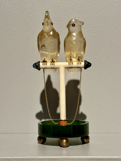 photo of the object on display at museum “Perched on a T-shaped ivory stand is a pair of topaz cockatoos with ruby eyes and gold feet. They are connected by gold chains to a pink enamel and gold mount set in a nephrite base supported by four gold ball feet. Carl Fabergé was particularly fond of birds and kept a pet cockatoo. Such hardstone animals were a popular part of Faberge's output in the early decades of the 20th century. Inspired by Japanese netsuke, which Fabergé collected in great numbers, these tiny animals have a life-like quality. Fabergé was even commissioned to make portraits of specific pets, most famously for King Edward VII and his wife Queen Alexandra.” https://art.thewalters.org/object/57.1913/