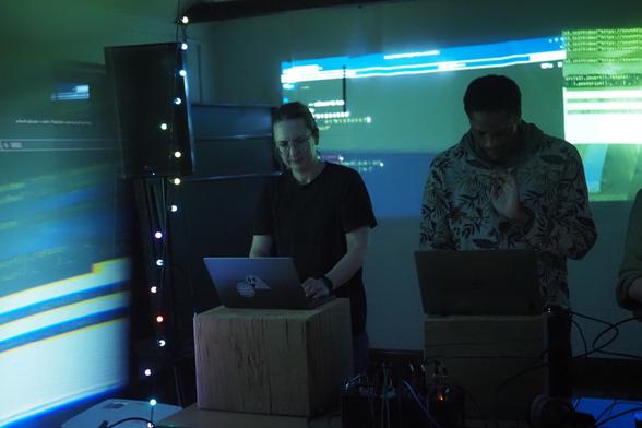 Two people doing live coding at their laptops with a projection behind them. The image is distorted slightly due to the use of a triangular prism